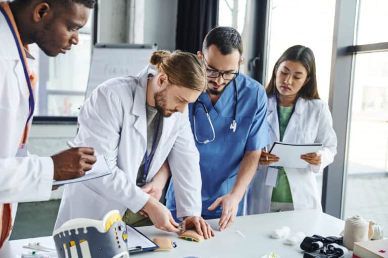 young student making injection in training pad near medical equipment, healthcare worker and multiethnic team in white coats during first aid seminar, skills development concept